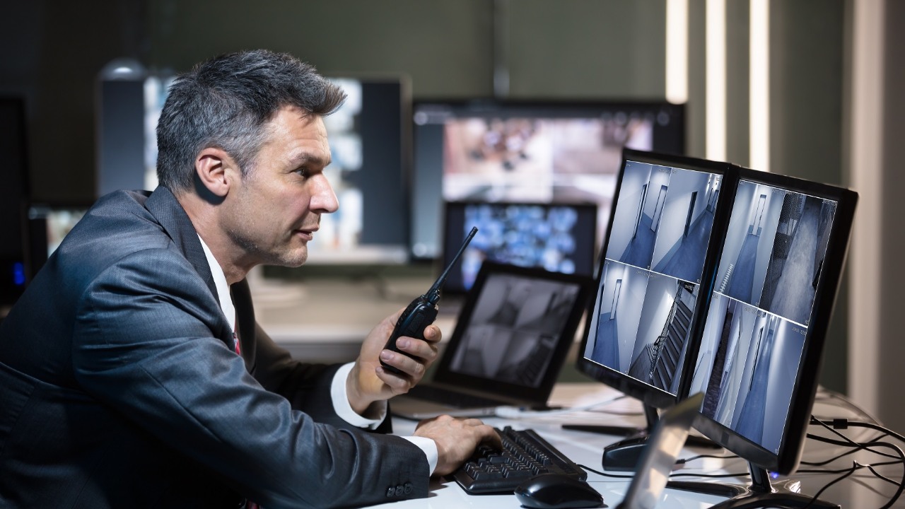 Businessman Talking On Walkie Talkie While Looking At CCTV Camera Footage On Multiple Computer Screen